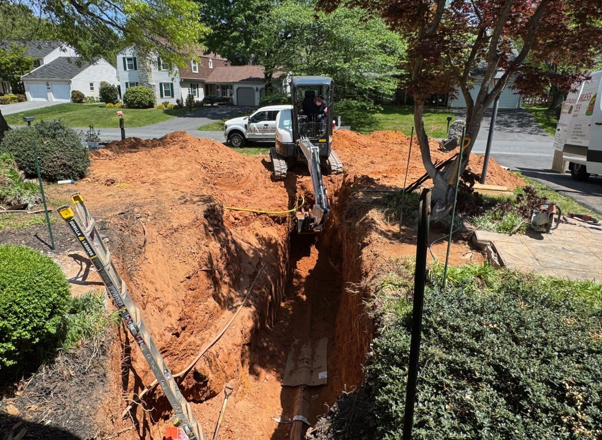 Deep sewer line trench across a front yard with a mini excavator and shoring ladder